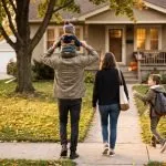 A father carrying a toddler on his shoulders walks with his wife and young son toward their house on an autumn afternoon